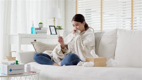 Sick Young Asian Woman Sit Under Blanket On Sofa And Sneeze With Tissue