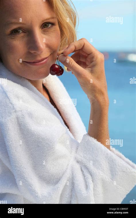 50 Years Old Blonde Woman Dressed In Bathrobe In Front Of The Sea Taking Cherries In Her Fingers
