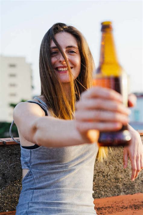 Beautiful Lively Smiling Young Female Holding Beer Bottle In Stretched Out Hand And Looking At