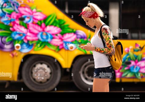 A Blonde Woman With A Phone In Hand Is Standing Against The Background Of A Yellow Bus Modern