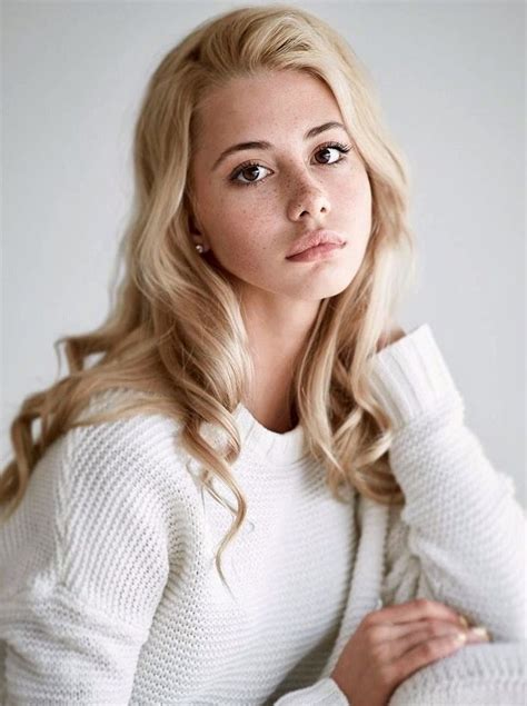 A Woman With Long Blonde Hair Wearing A White Sweater And Looking At The Camera While Leaning On