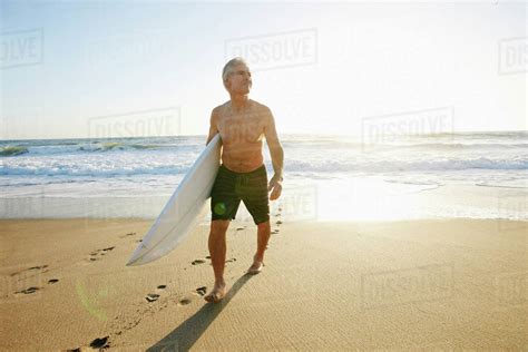 Older Caucasian Man Walking On Beach Carrying Surfboard Stock Photo