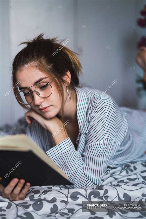 Portrait Of Brunette Girl In Glasses Reading Book And Lying On Bed