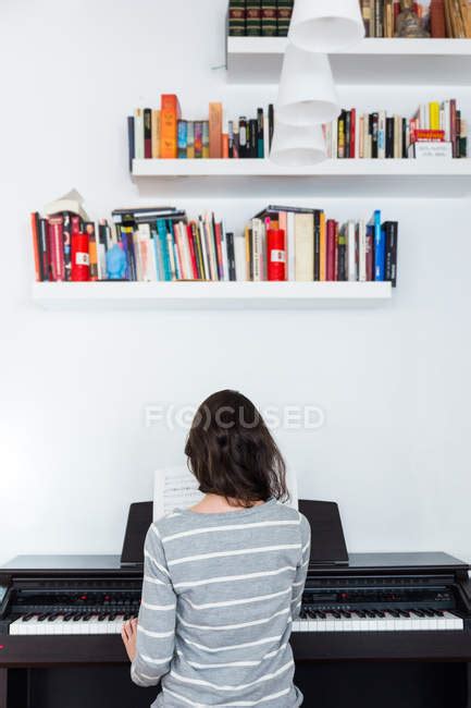 Rear View Of Girl Playing Piano Near Wall With Book Shelves — Brunette