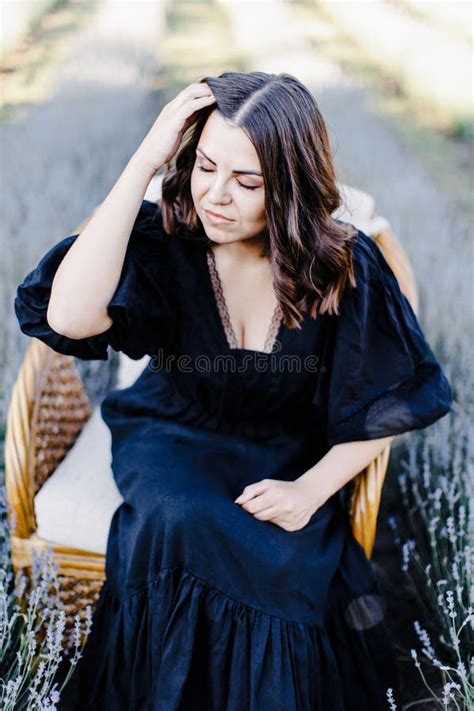 Attractive Young Woman In Black Dress And Sitting In Chair Surrounded By Lavender Field Stock
