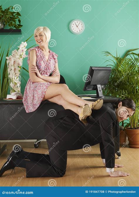 Female Office Worker Resting Legs On Man Kneeling On Hands And Knees In Office Portrait Stock