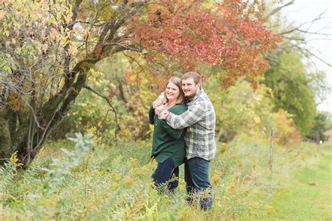 In Green And Grey Plaid An Engaged Couple Smile In Front Of A Red Fall Tree Engagement Season
