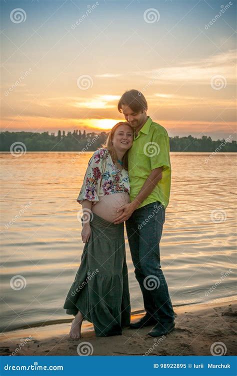 A Pregnant Beautiful Woman With Her Husband On The River Bank Beach Smiling And Touching Her