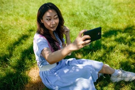 attractive asian girl taking a selfie on smart phone sitting in the park on the grass stock