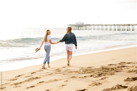Two Friends Holding Hands And Walking On The Beach With Pier And Ocean