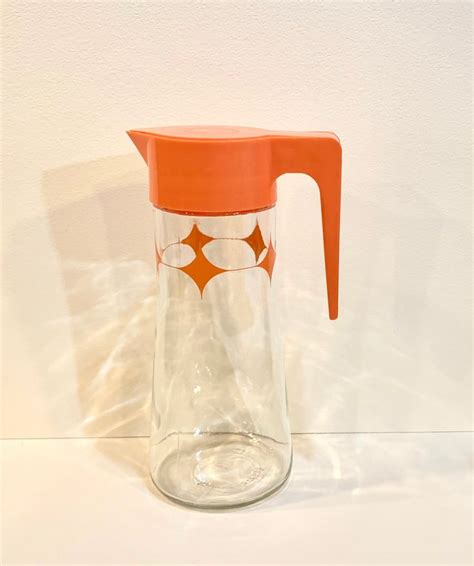 A Glass Pitcher With An Orange Lid On A White Surface Next To A Wall