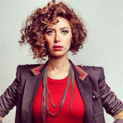A Woman With Curly Hair Wearing A Red Top And Black Jacket Is Posing For The Camera