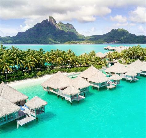 An Aerial View Of Some Huts In The Water With Palm Trees And Mountains