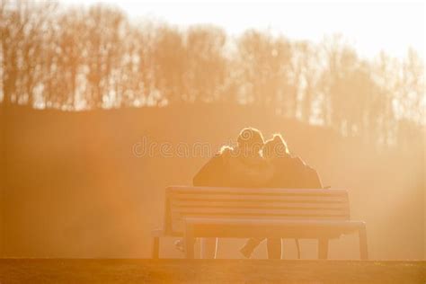 Lovely Couple Sit On Bench In Park Lighted By Beautiful Sunset