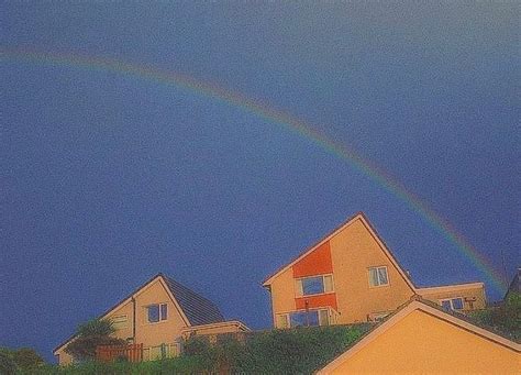 A Rainbow Shines In The Sky Over Houses On A Hill With Trees And Bushes