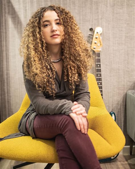 A Woman With Curly Hair Sitting On A Yellow Chair In Front Of A Guitar Case