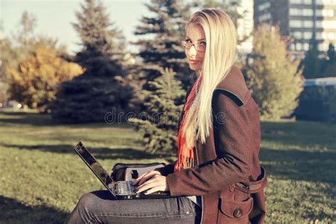 Side View Of Happy Blonde Woman In Eyeglasses Sitting On Bench In Park With Laptop Computer