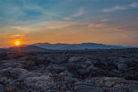 The Sun Is Setting Over Rocky Terrain With Mountains In The Background