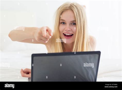 Happy Young Woman Pointing Her Finger Towards The Camera While Using A Laptop Computer In Bed