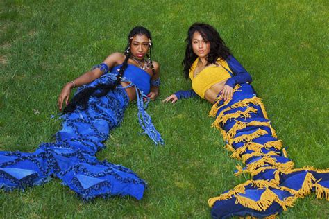 Two Women In Mermaid Costumes In A Green Outdoor Garden Laying On The Grass Next To Each Other