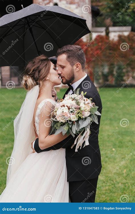 Gorgeous Bride And Stylish Groom Passionately Kissing Under Umbrella In