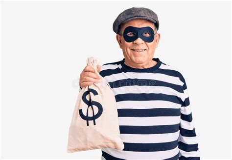 senior handsome man wearing burglar mask holding money bag looking positive and happy standing