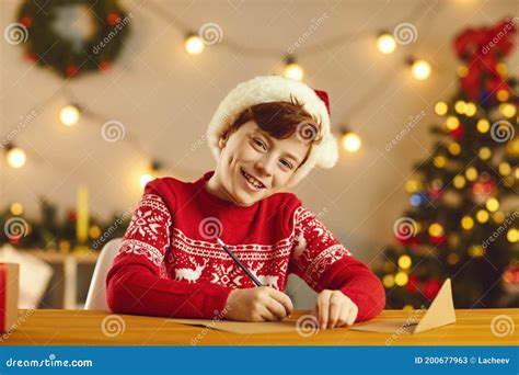 Smiling Boy In Red Hat And Sweater Sitting And Writing Letter To Santa